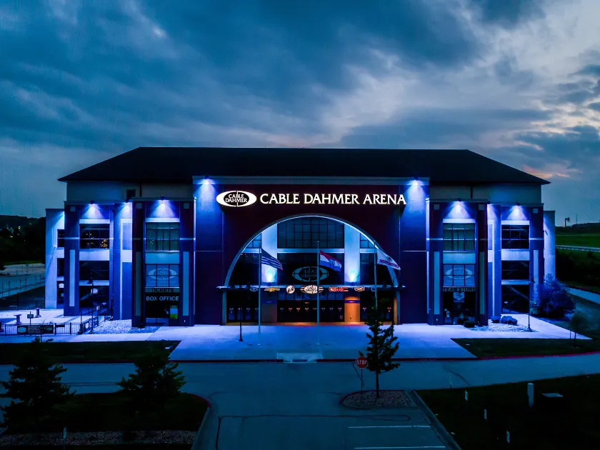 Entrada principal de Cable Dahmer Arena al atardecer, con la fachada iluminada en azul, un letrero iluminado de CABLE DAHMER ARENA sobre las puertas de la taquilla y banderas al frente — Independence Community Ice (Cable Dahmer Arena)