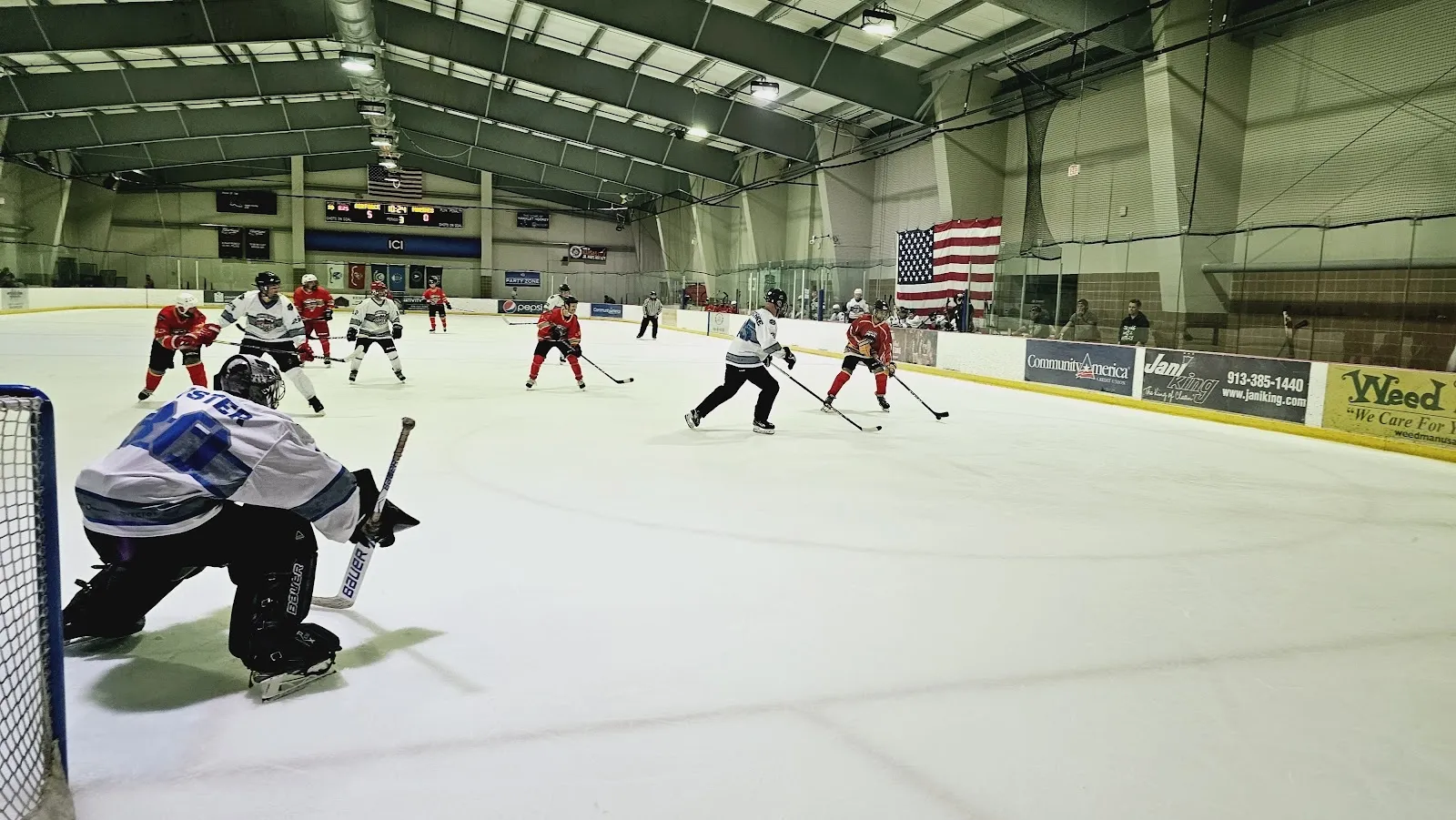 Un partido en el extremo lejano de una pista amplia, con jugadores repartidos por el hielo detrás de un portero en el borde izquierdo, bajo un techo de acero verde con una bandera de Estados Unidos en la pared del extremo. — Independence Community Ice (Cable Dahmer Arena)