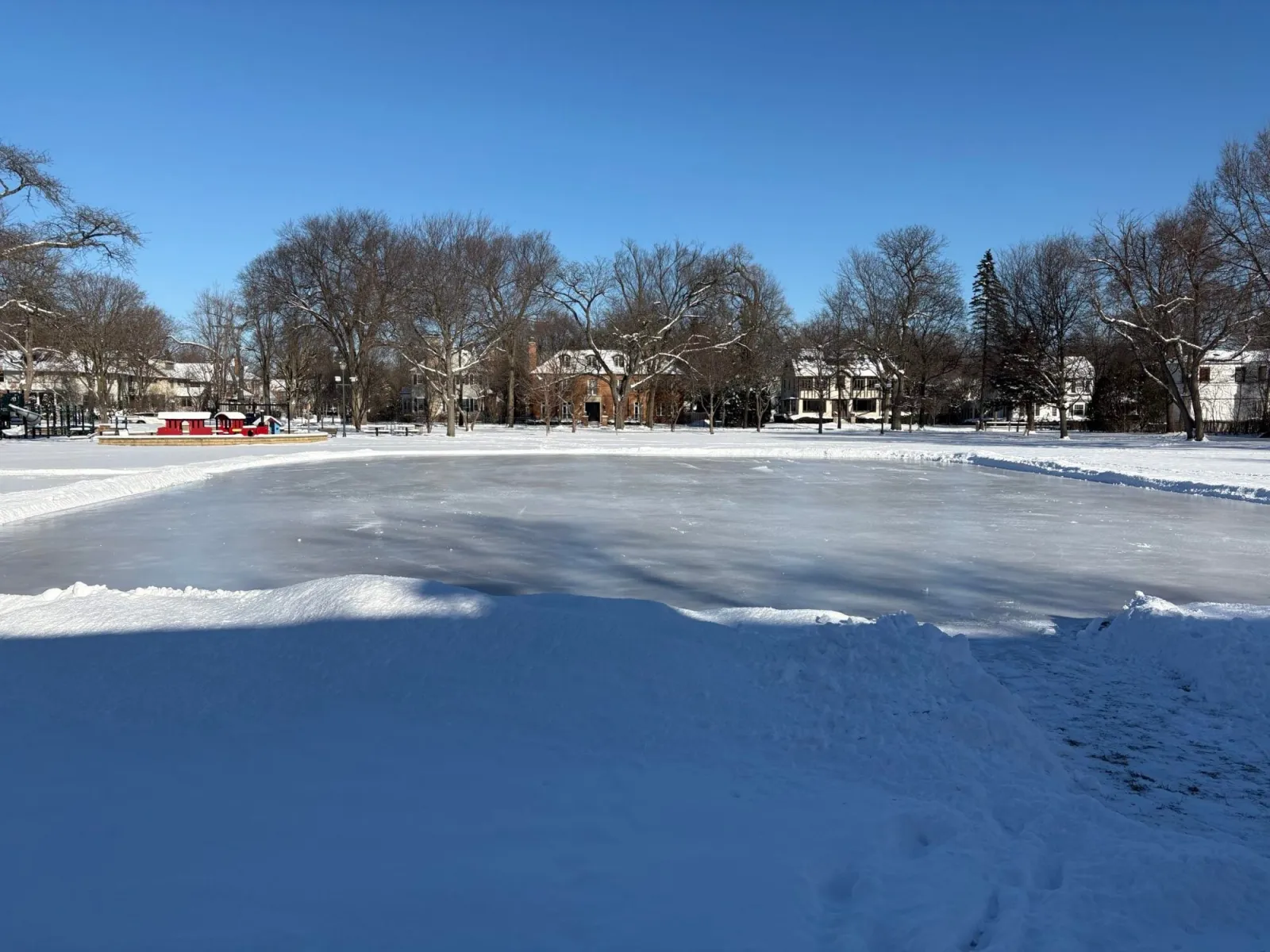 La pista de hielo exterior de Indian Hill Park, en Winnetka, en invierno: una superficie helada blanca con bancos de nieve en el borde y árboles sin hojas y casas al fondo. — Indian Hill Park Outdoor Ice Rink