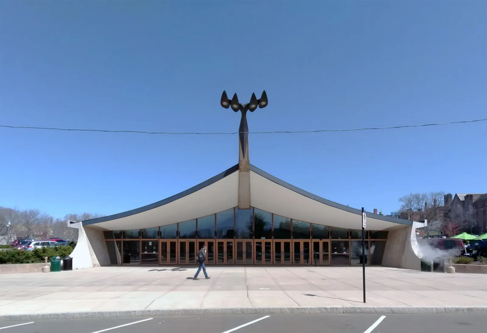 La fachada frontal de la pista: el techo envolvente de concreto y cobre, levantado al centro sobre su columna vertebral, una pared de entrada de vidrio y madera debajo y un mástil de tres lámparas arriba. — Ingalls Rink