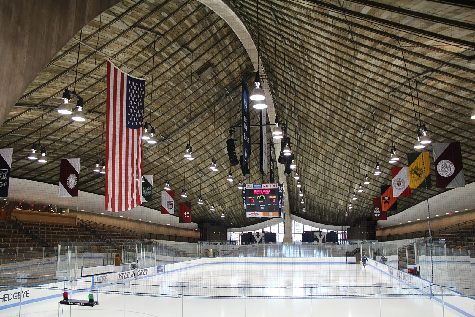 El hielo vacío visto desde un extremo, con el techo curvo de madera de la pista arqueándose arriba, una bandera de Estados Unidos y banderines de escuelas de la Ivy League colgados de él y el marcador en el extremo del fondo. — Ingalls Rink
