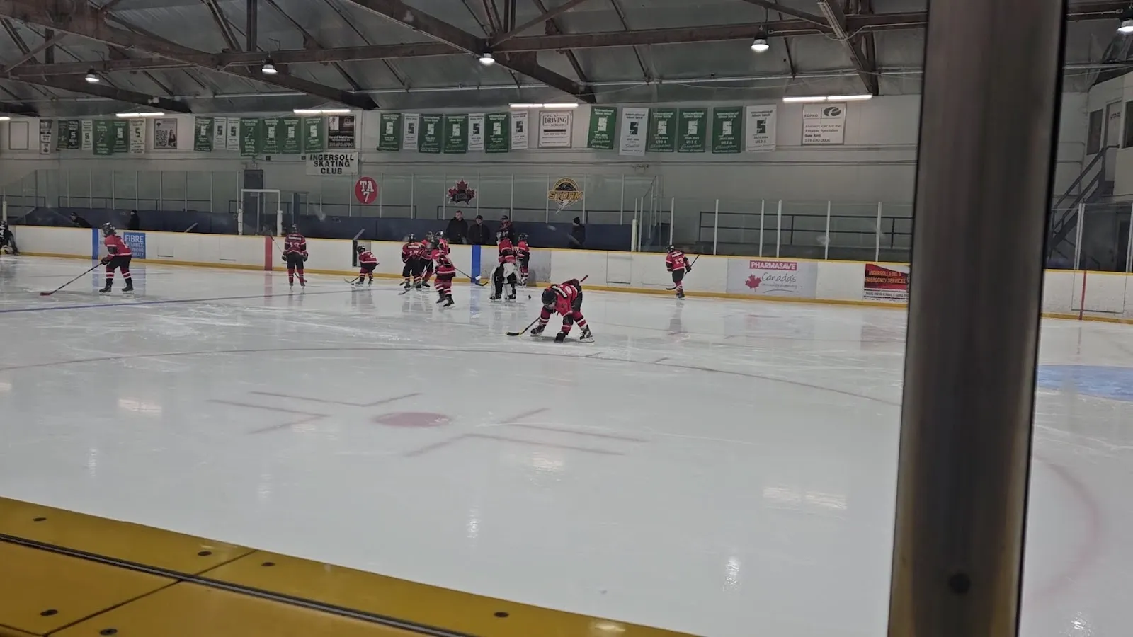 Un partido visto desde el barandal en un extremo de una pista amplia, con jugadores repartidos por el hielo blanco y una franja amarilla de vallas en el lado cercano. — Ingersoll District Memorial Centre