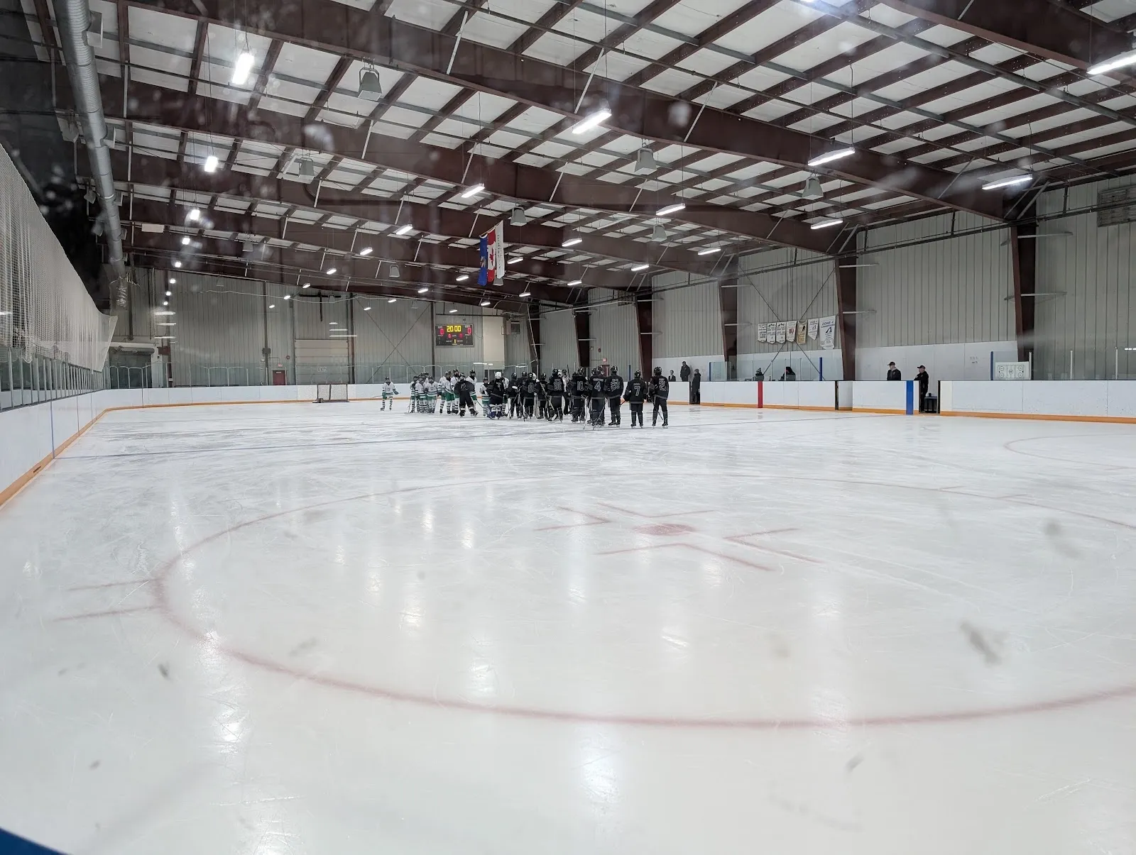 Un equipo alineado en las vallas del fondo de una pista amplia, con círculos y una línea roja sobre el hielo blanco bajo un oscuro techo de acero. — Innisfail Twin Arena