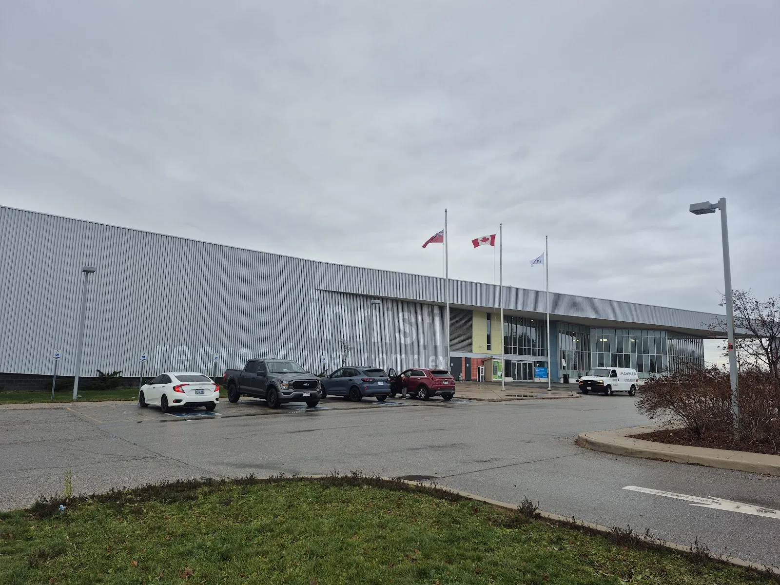 The long metal-clad Innisfil Recreational Complex with its lettering, flags and glass entrance, seen from the parking lot. — Innisfil Recreational Complex