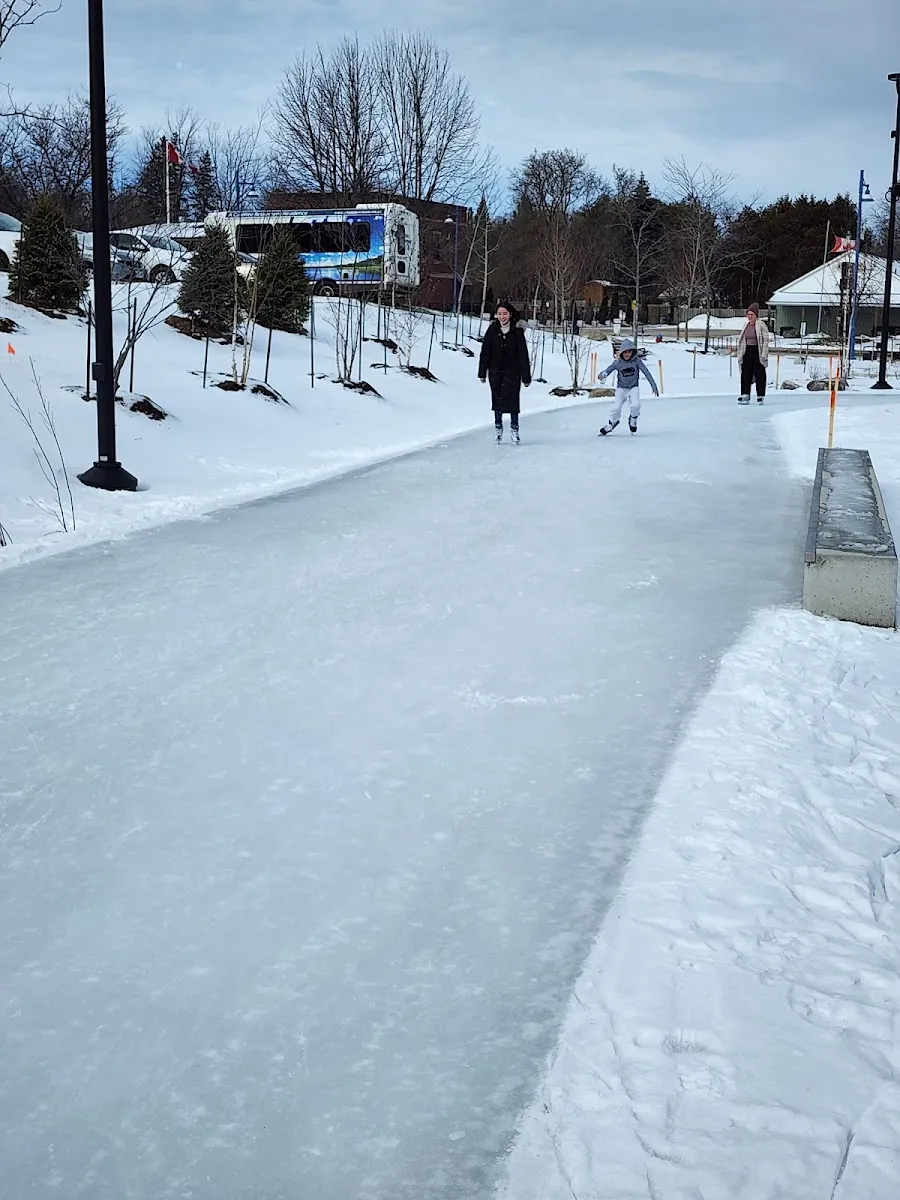 El hielo de la pista de patinaje de Innisfil Town Square en invierno, con unos cuantos patinadores deslizándose por el sendero entre bancos de nieve y postes de luz — Innisfil Town Square Skating Trail