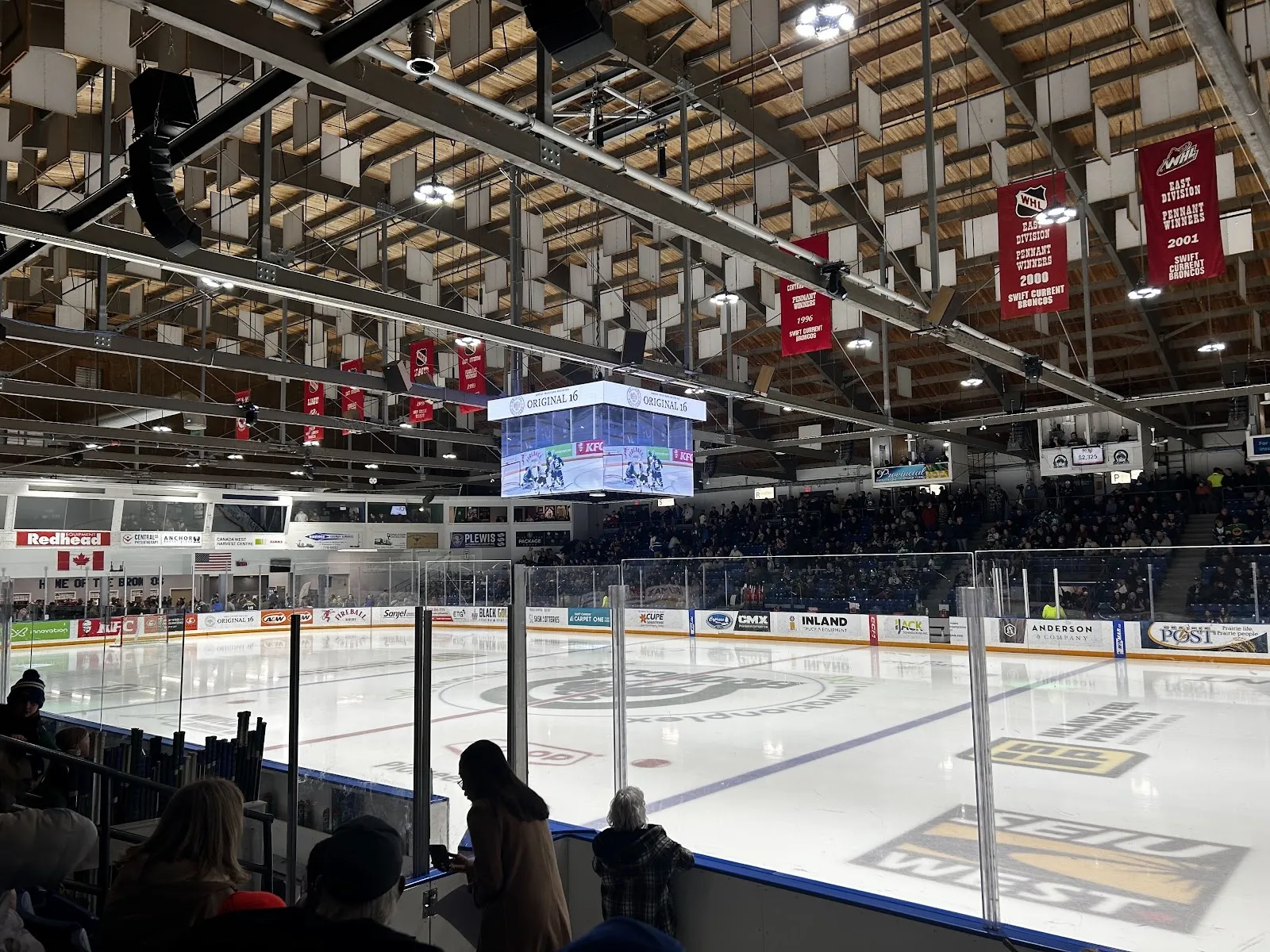 The arena bowl seen from the lower seats, players on the ice below a roof hung with banners and a video board over centre ice, spectators filling the stands. — InnovationPlex