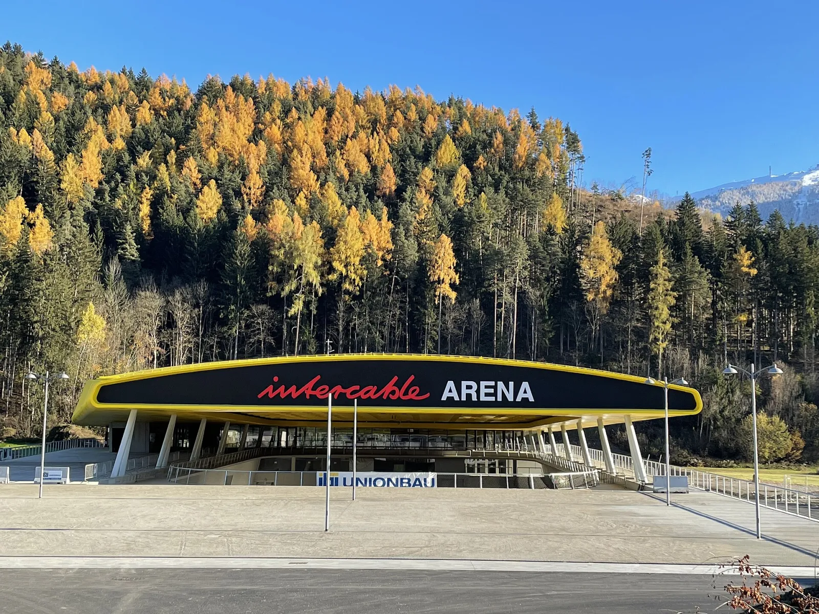L’Intercable Arena di Brunico vista dal piazzale, con il tetto dal bordo giallo che porta il nome dell’arena e il versante boscoso della collina dietro. – Intercable Arena Bruneck