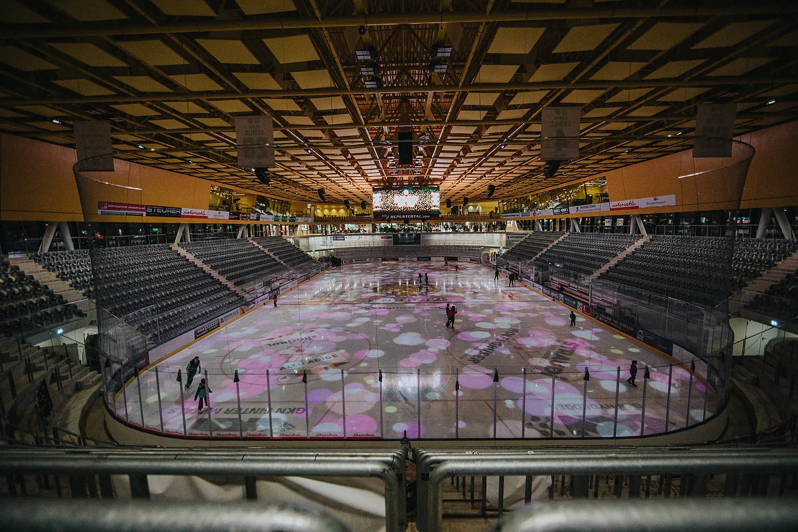 Il ghiaccio dell’Intercable Arena illuminato da proiezioni colorate durante un turno di Disco on Ice, visto dalle tribune con alcuni pattinatori sul ghiaccio. – Intercable Arena Bruneck