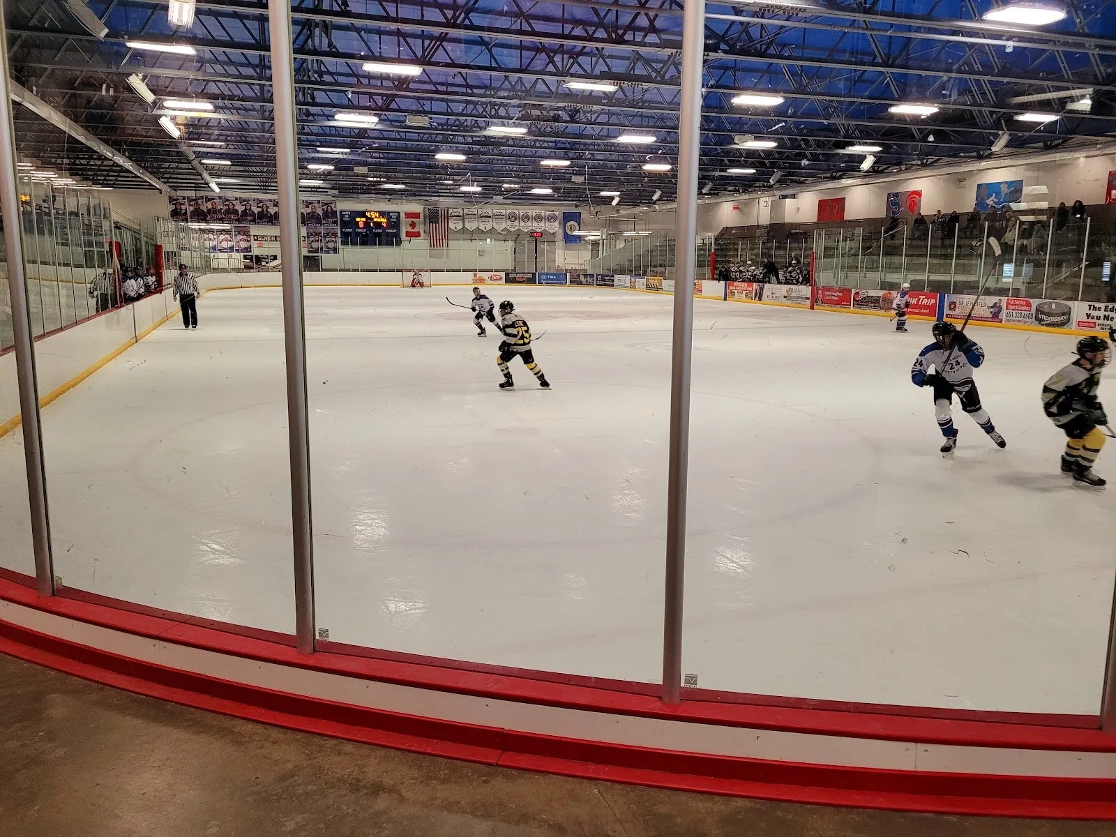 Un partido visto a través del vidrio de la pista y los zócalos rojos, con jugadores repartidos por una pista amplia bajo un techo de acero iluminado de azul. — Veterans Memorial Community Center Ice Arenas