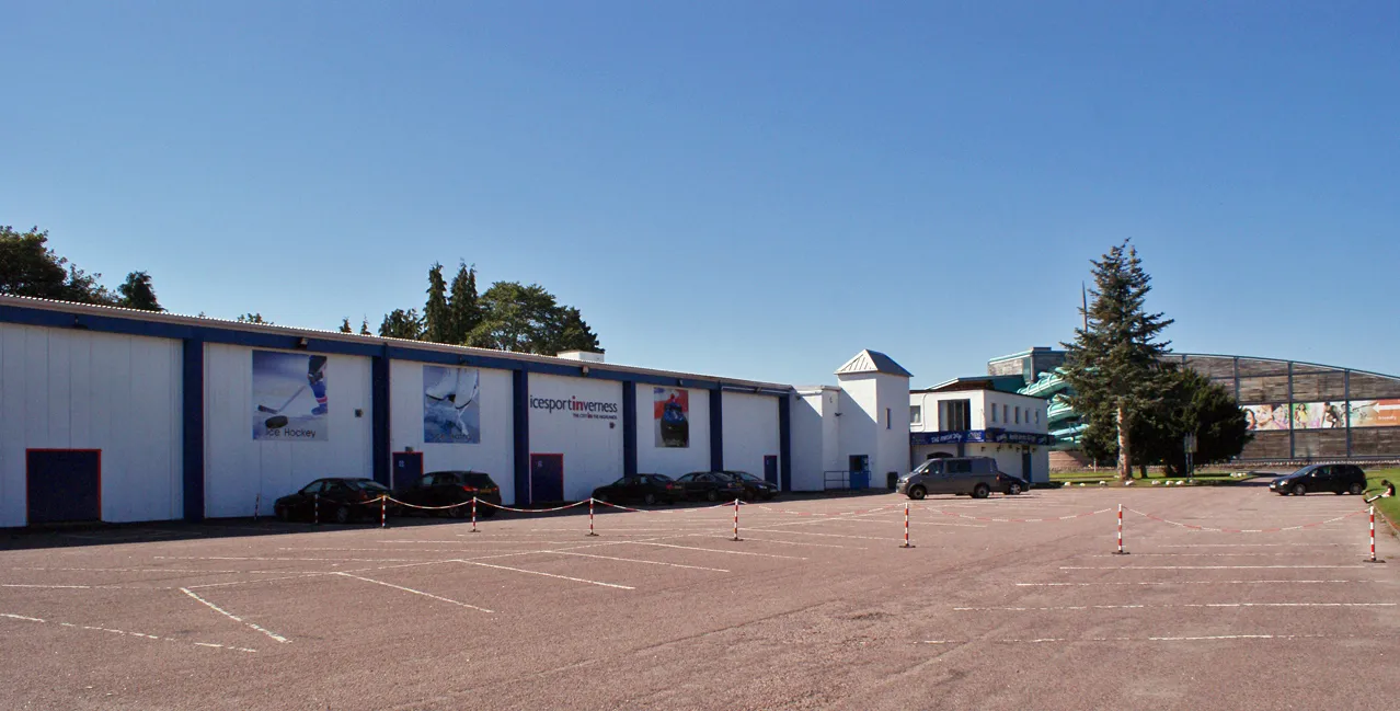 The long white side of the ice centre with its ice hockey, skating and curling posters, seen across the car park. — Inverness Ice Centre