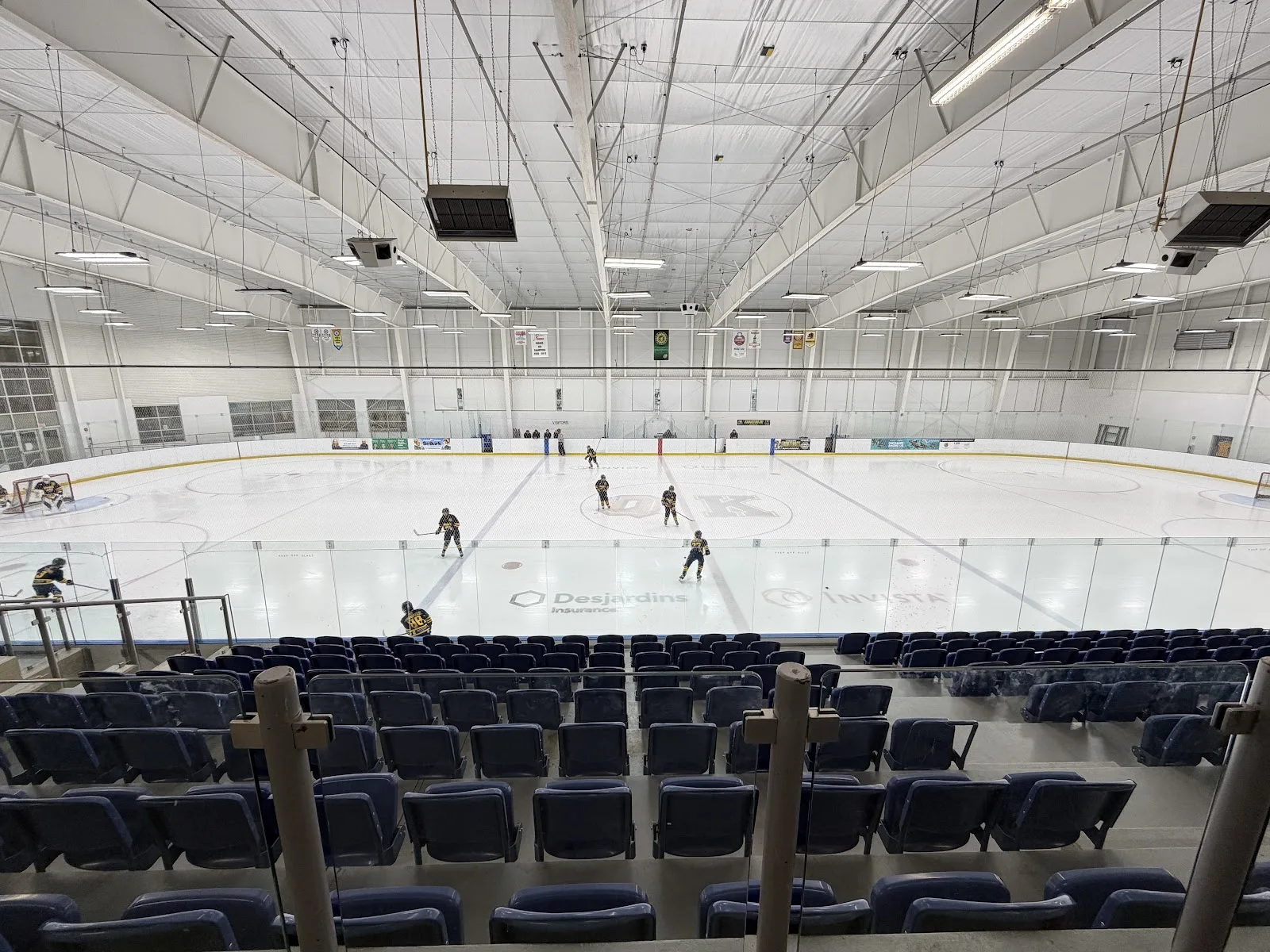 The Desjardins Insurance Rink seen from the upper rows of blue seats, with a few players at centre ice under the white roof trusses. — INVISTA Centre