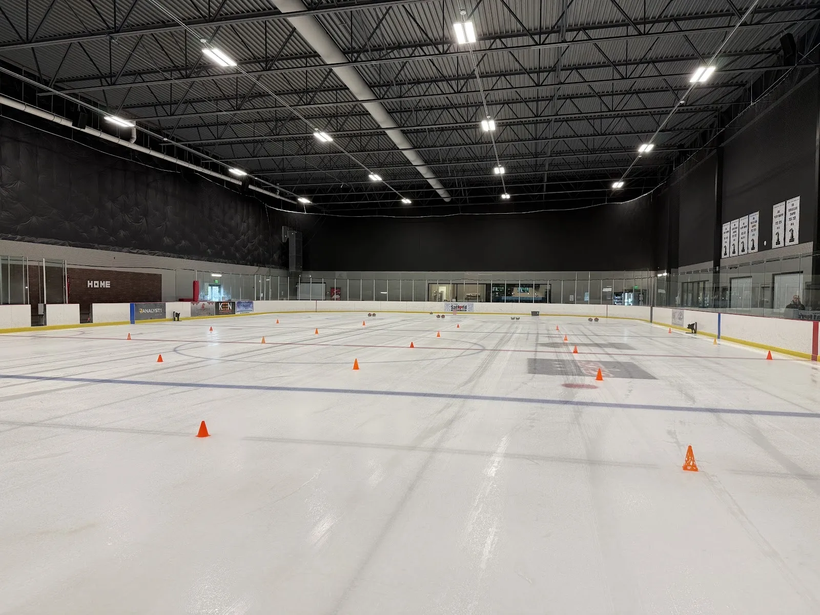 A wide empty sheet with a few orange cones on the ice, a high dark steel ceiling and the boards behind it. — Ion International Training Center (Leesburg)