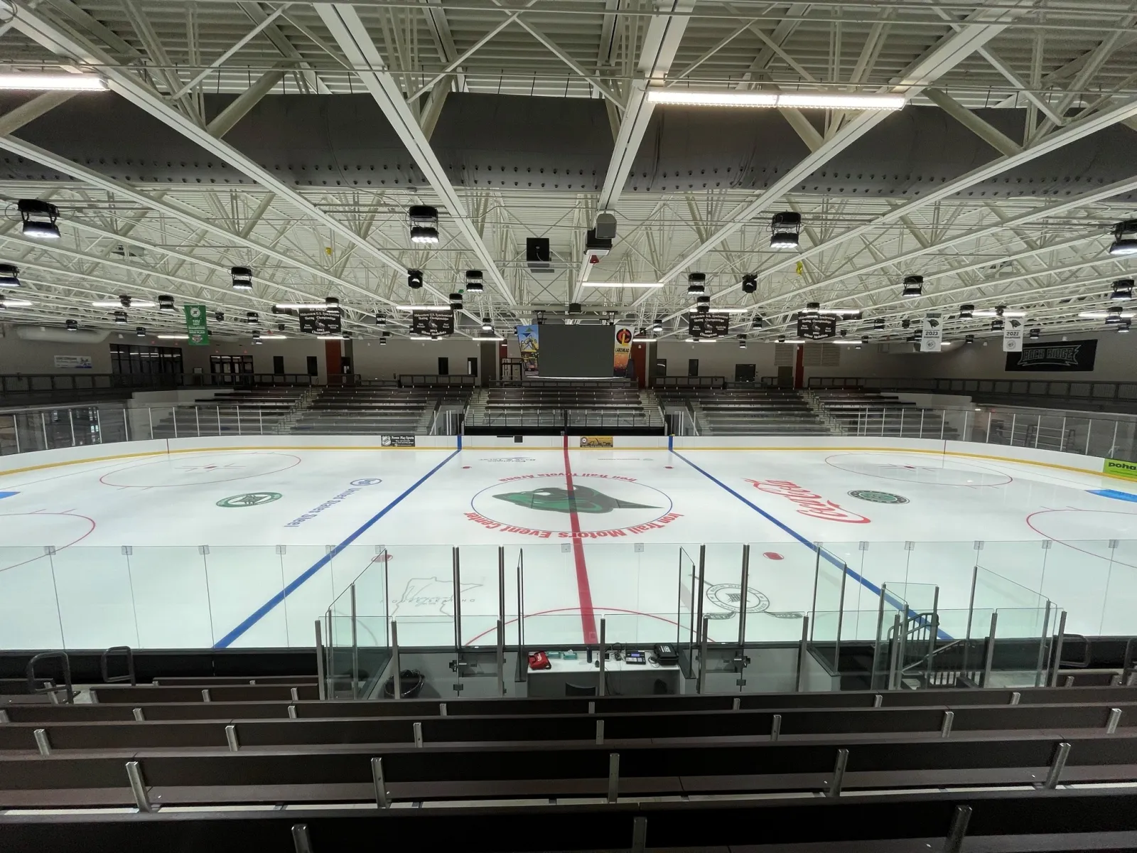 Empty ice sheet and bleachers seen from an elevated corner, with "Iron Trail Motors Event Center" lettered in the center-ice logo and a Rock Ridge banner above the scoreboard. — Iron Trail Motors Event Center