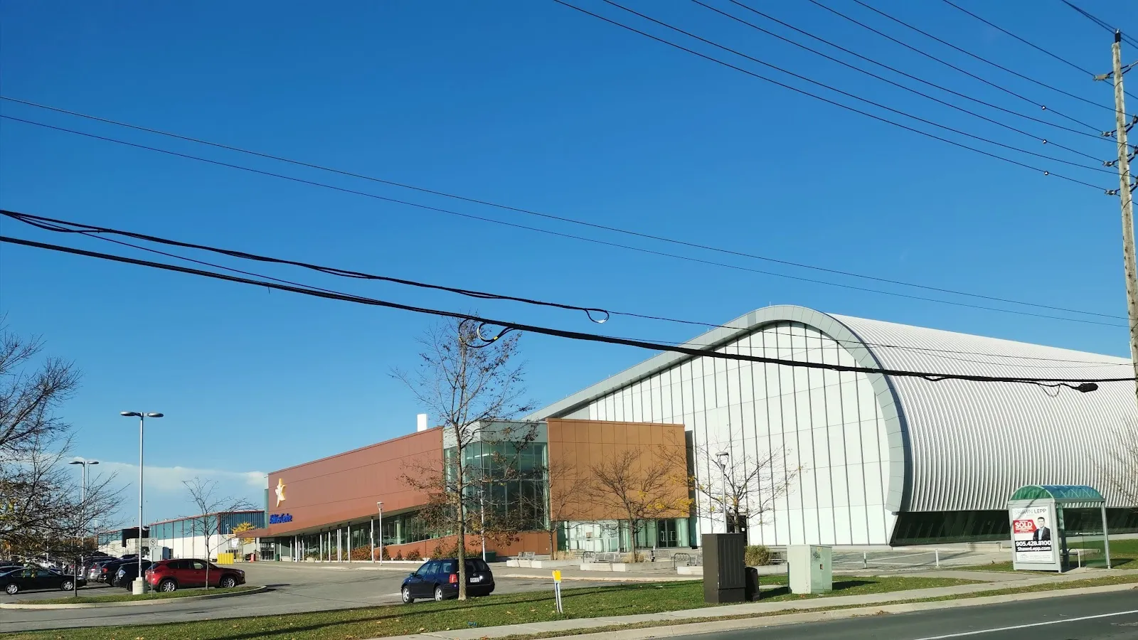 El Iroquois Park Sports Centre en Whitby desde Victoria Street, con su techo blanco curvo y su frente de ladrillo y vidrio bajo un cielo despejado — Iroquois Park Sports Centre
