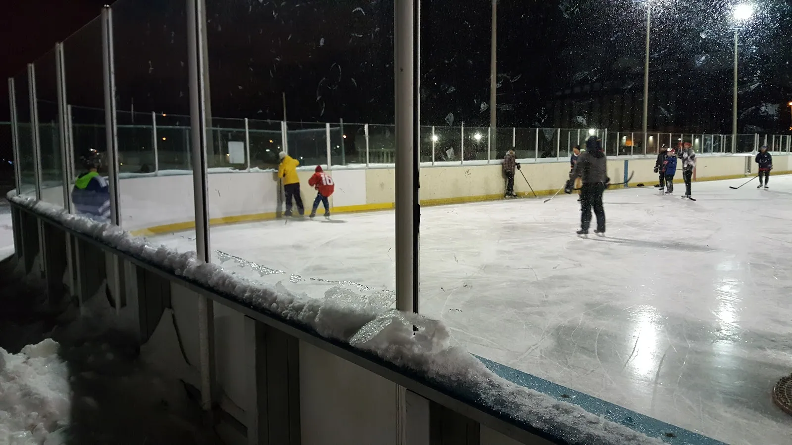 Patinadores y jugadores de hockey en la pista al aire libre Irving W. Chapley de noche, vistos a través de las vallas, con reflectores y una cerca detrás — Irving W. Chapley Community Centre Outdoor Ice Rink