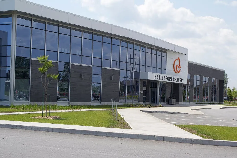 La façade de verre et de brique foncée du complexe, avec l’enseigne ISATIS SPORT CHAMBLY au-dessus de l’entrée et une pelouse à l’avant. — Isatis Sport Chambly