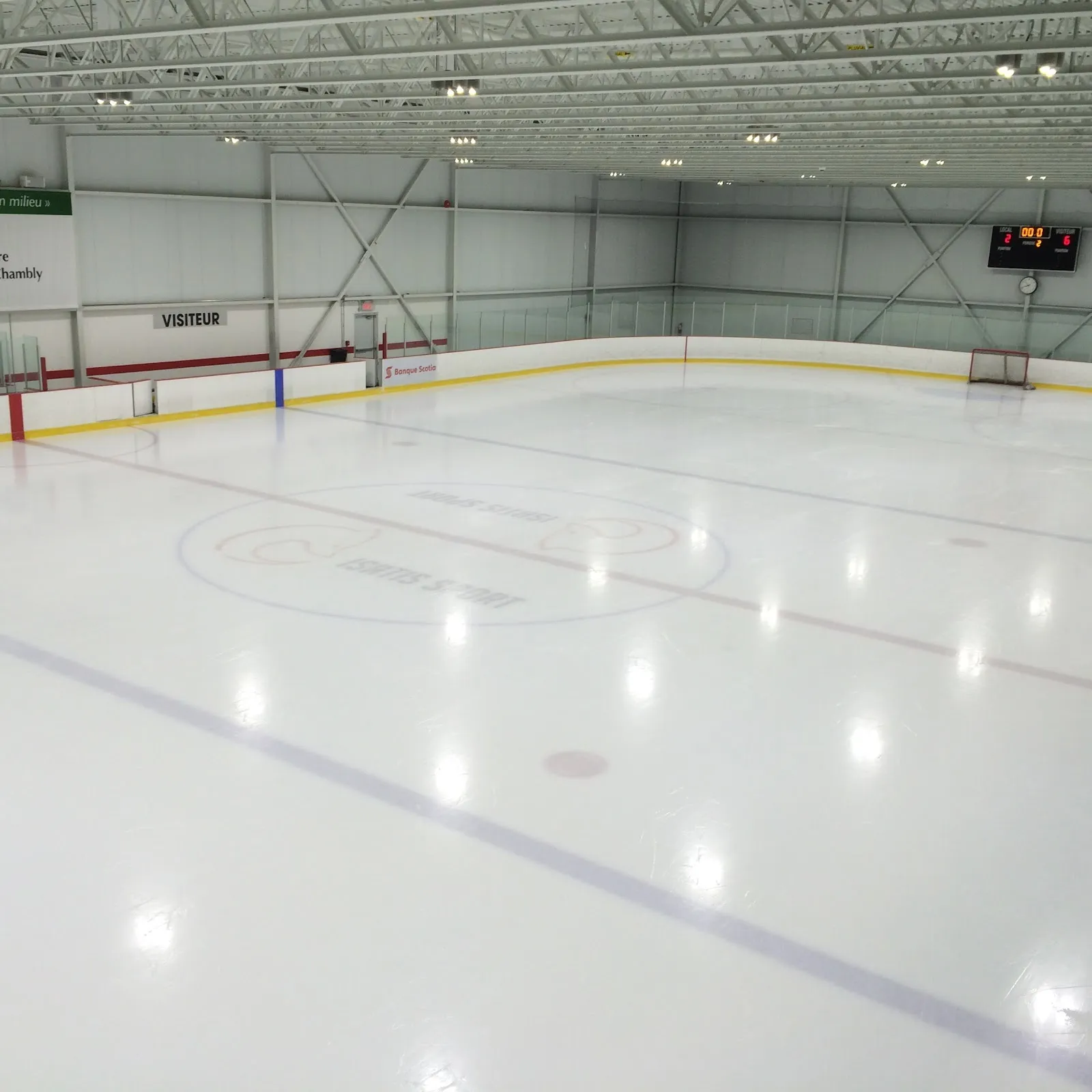 Une grande surface de glace vide avec une ligne bleue et un cercle de mise au jeu, un haut plafond garni de lumières au-dessus des bandes. — Isatis Sport Chambly