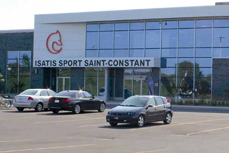 The glass-fronted entrance of the Isatis Sport Saint-Constant complex with its name across the facade and cars parked in front. — Isatis Sport Saint-Constant