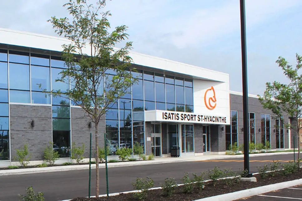 The long glass and grey-panel front of the complex, signed Isatis Sport St-Hyacinthe beside an orange fox logo, with young trees along the parking lot. — Isatis Sport St-Hyacinthe