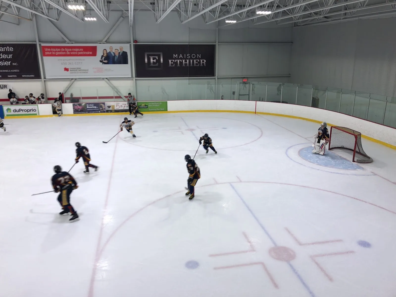 A wide view along the boards of a bright sheet, players spread across the ice around a faceoff dot under a long ceiling. — Isatis Sport St-Hyacinthe