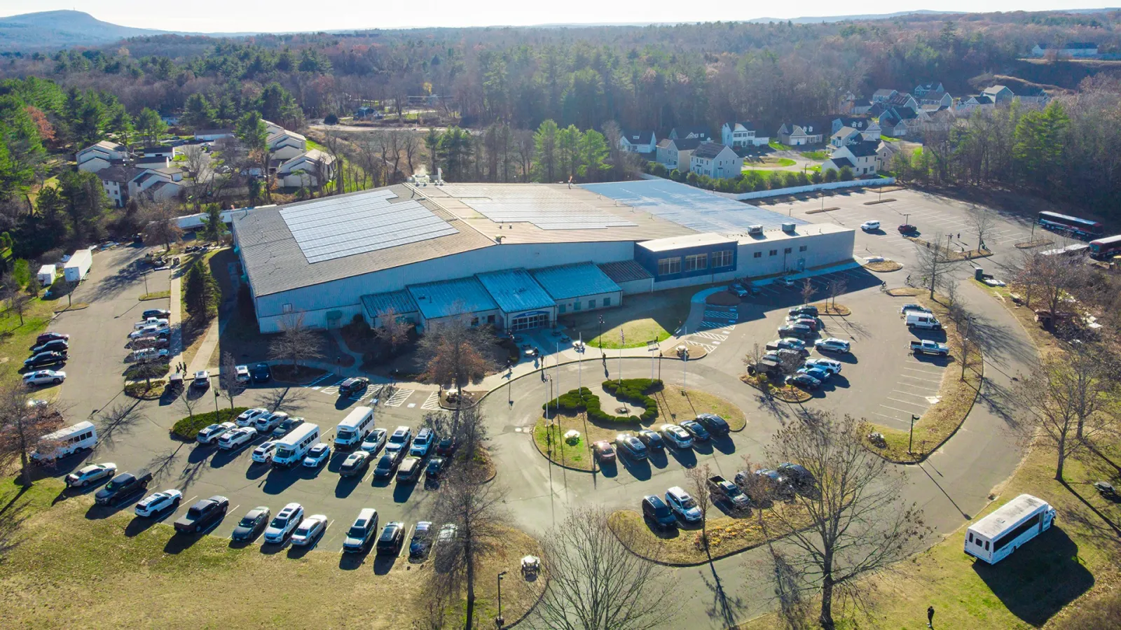 Vista aérea del gran edificio blanco del International Skating Center of Connecticut con techo de paneles solares, rodeado de estacionamiento y con una glorieta con asta de bandera en la entrada, en Simsbury, CT. — International Skating Center of Connecticut