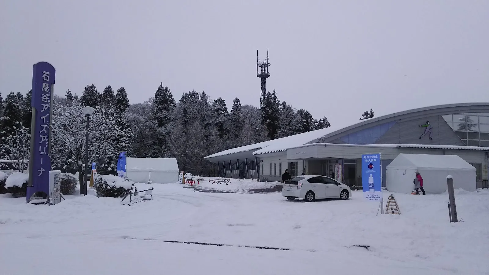 降雪のあとの、曲線の屋根をもつ石鳥谷アイスアリーナの建物。入口の左に名称の看板があり、後ろに木立が見えます。 — Ishidoriya Ice Arena