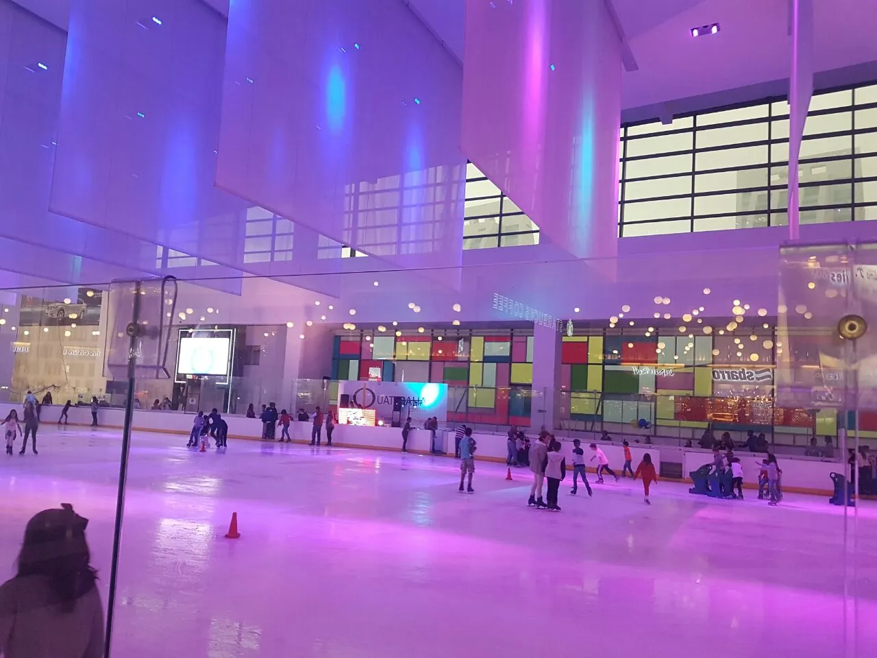 Pista de hielo en el atrio de un centro comercial con iluminación morada, patinadores por toda la pista y locales comerciales detrás de las vallas del fondo. — ISK8 Interlomas