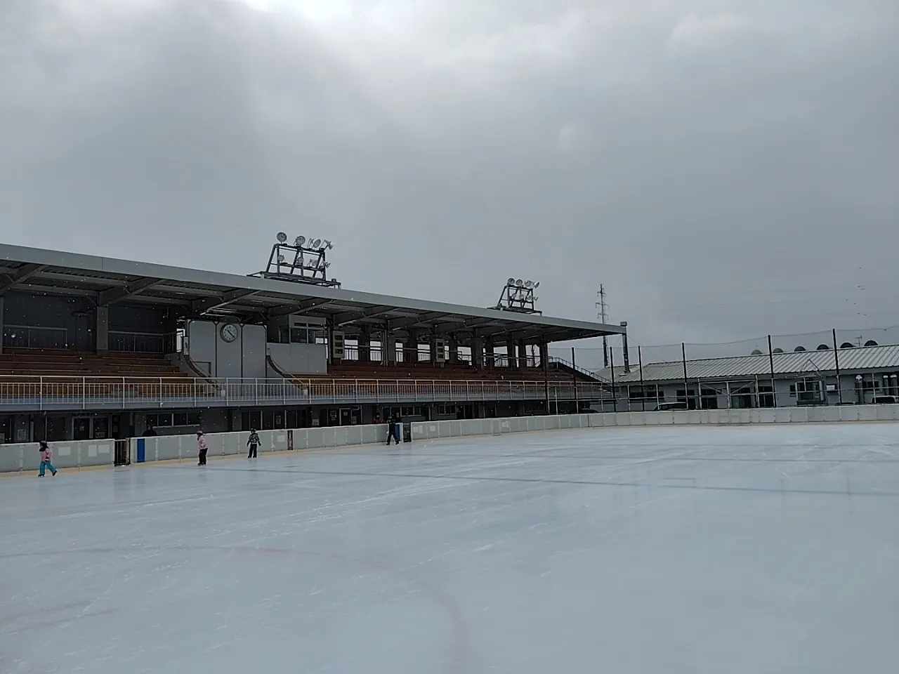 The outdoor ice of the Iwate Prefectural Skating Rink under a grey winter sky, with the covered grandstand along one side and three skaters far off. — Iwate Prefectural Skating Rink