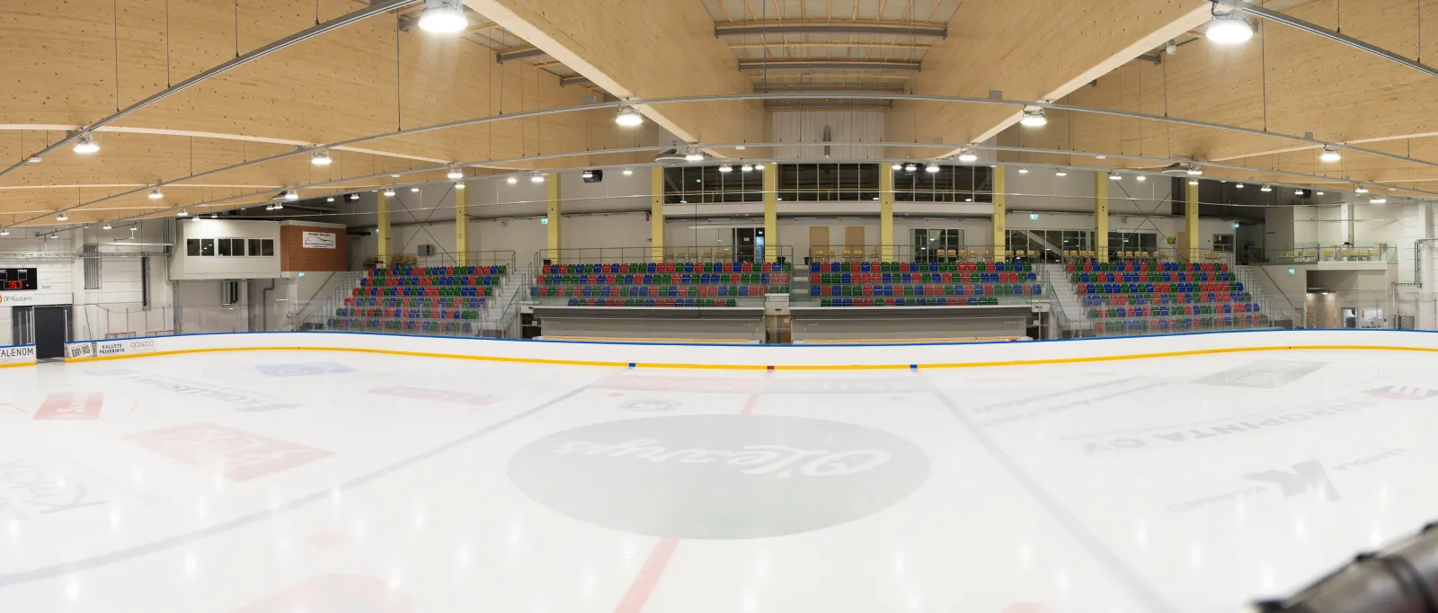 Panoramic view across the white ice of the Kuusamo EVO-areena from the rink, with the colourful grandstand seating and the wooden roof beams above. — Jäähalli EVO-areena