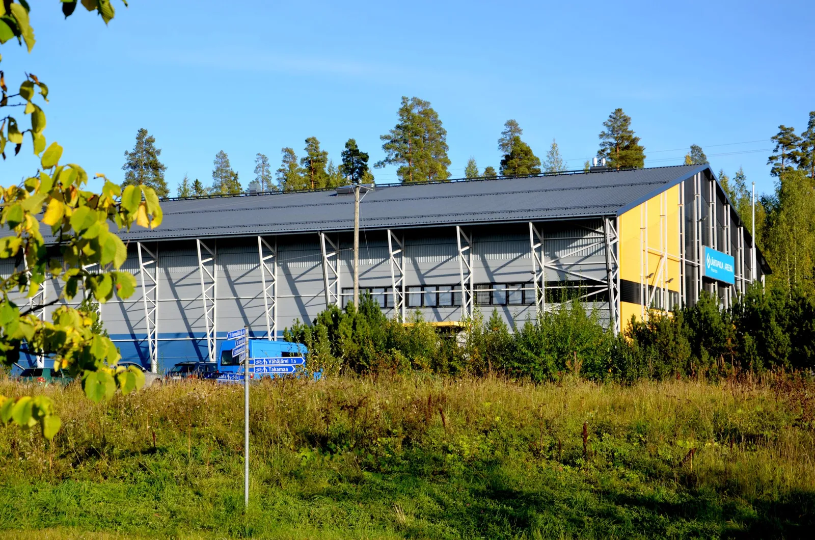 The Pirkkala ice hall seen across a grass field in summer: a long grey steel-framed hall with a yellow entrance end and pine trees behind. — Jäähallin ulkojää (Pirkkala)