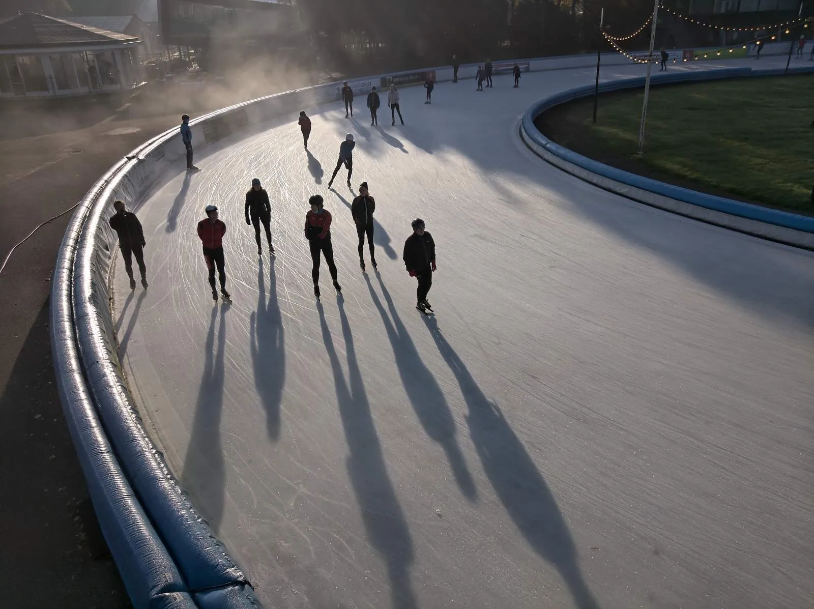 Skaters on the Jaap Eden outdoor ice track in low winter sun, long shadows stretching across the curve of the oval. — Jaap Eden IJsbaan