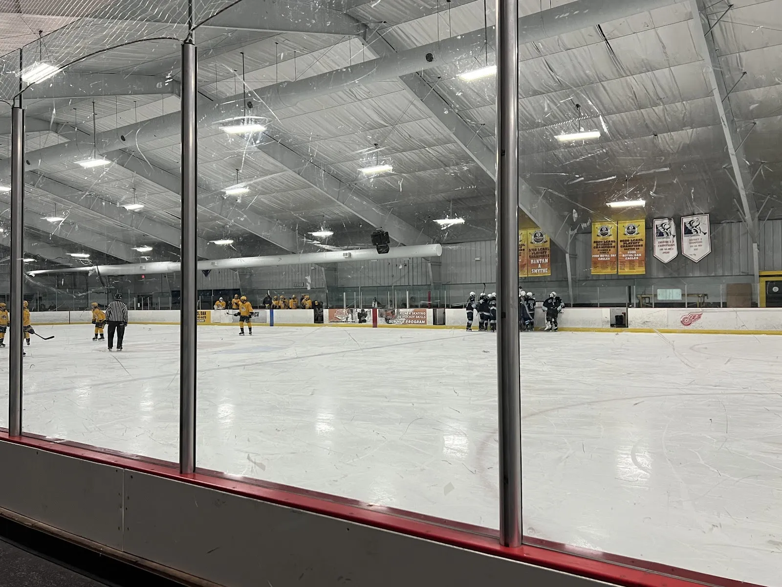 Empty ice sheet inside the arena with the glass and rail boards, a grey steel roof with lights, and championship banners on the far wall. — Jack Adams Ice Arena