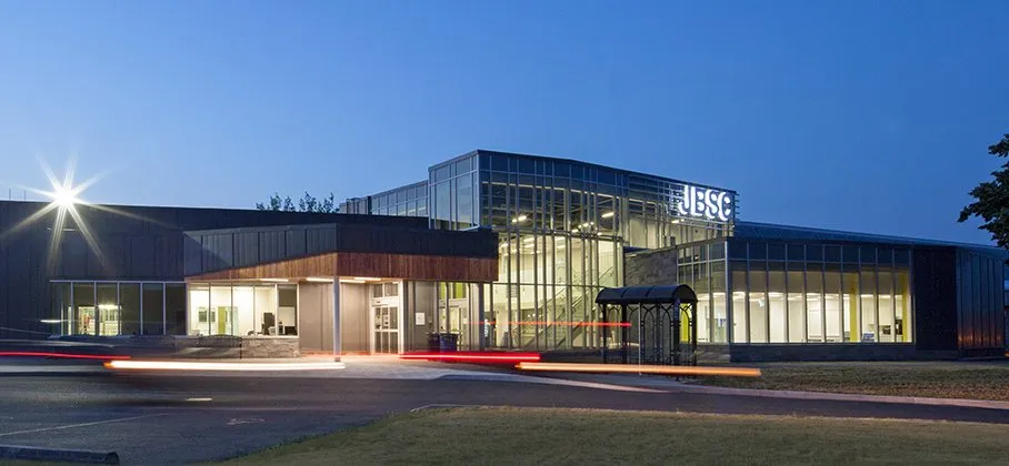 El complejo renovado al anochecer: un vestíbulo con fachada de vidrio y el letrero JBSC, y detrás el ala de la arena con revestimiento oscuro. — Jack Burger Sports Complex (Port Hope)