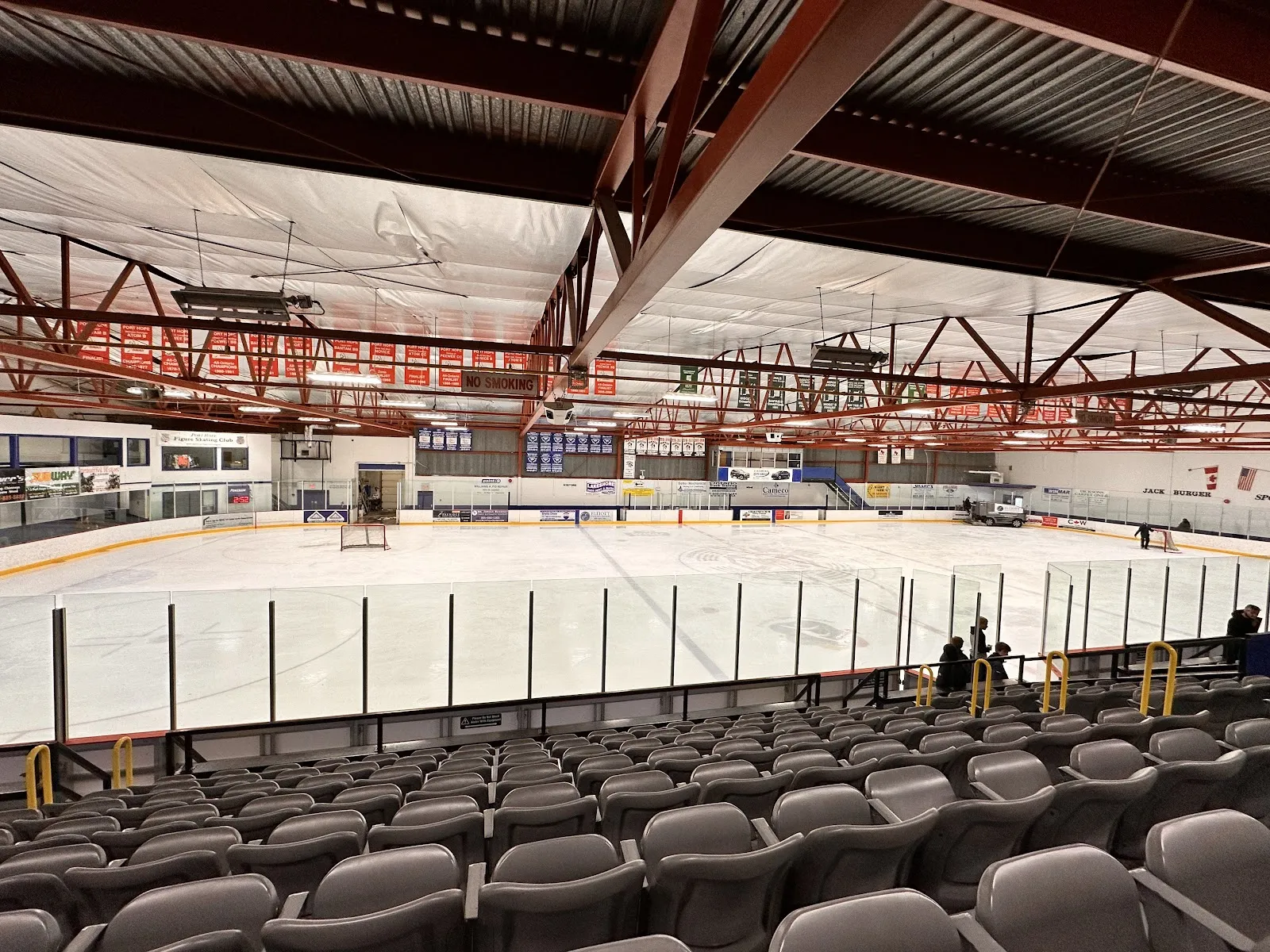 La pista desde los asientos grises: un techo de armaduras de acero rojas sobre el hielo, con mantas y el marcador en el extremo del fondo. — Jack Burger Sports Complex (Port Hope)