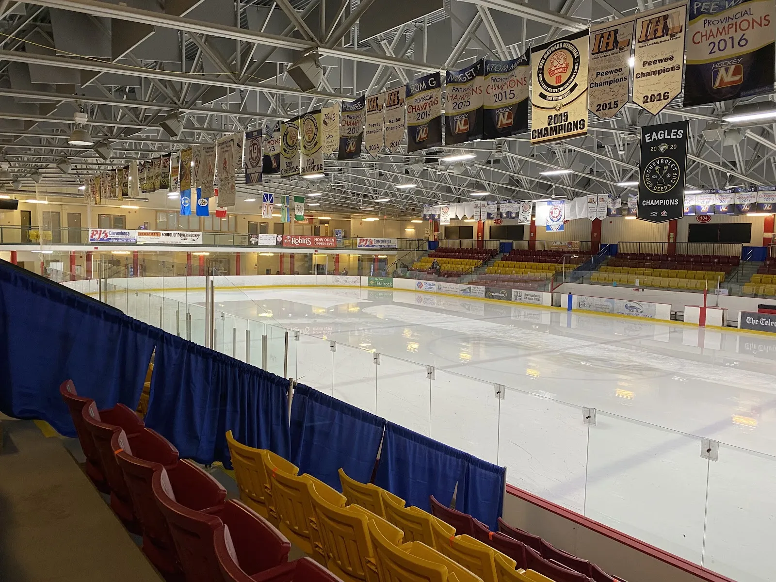 Toda la pista vista desde la tribuna superior, con asientos rojos y dorados de la arena en primer plano y mantas de campeonato (Hockey NL, campeones provinciales, Eagles) colgadas de las armaduras del techo. — Jack Byrne Regional Sport & Entertainment Centre