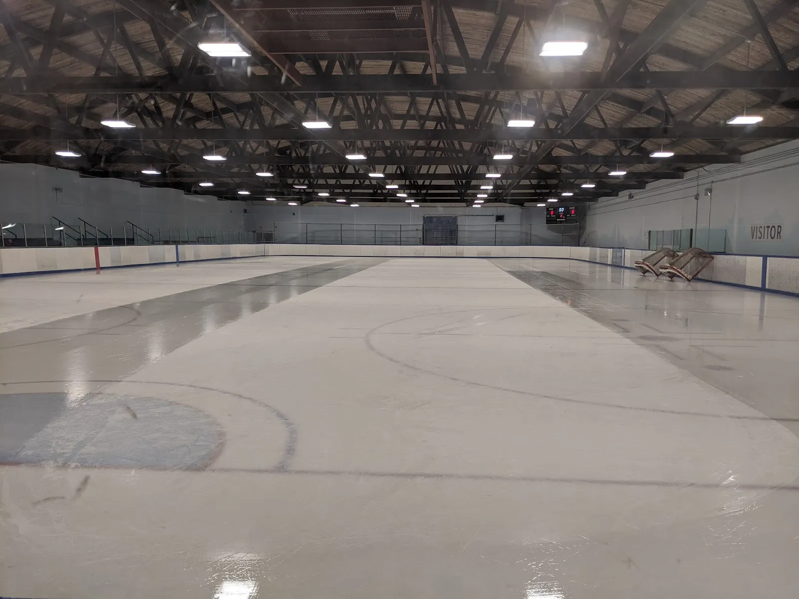 El hielo vacío de la Jack Hamilton Arena visto desde detrás del vidrio: hielo blanco recién preparado con las marcas de hockey, un techo de armaduras de acero oscuro con hileras de luces y un marcador en el extremo del fondo. — Jack Hamilton Arena