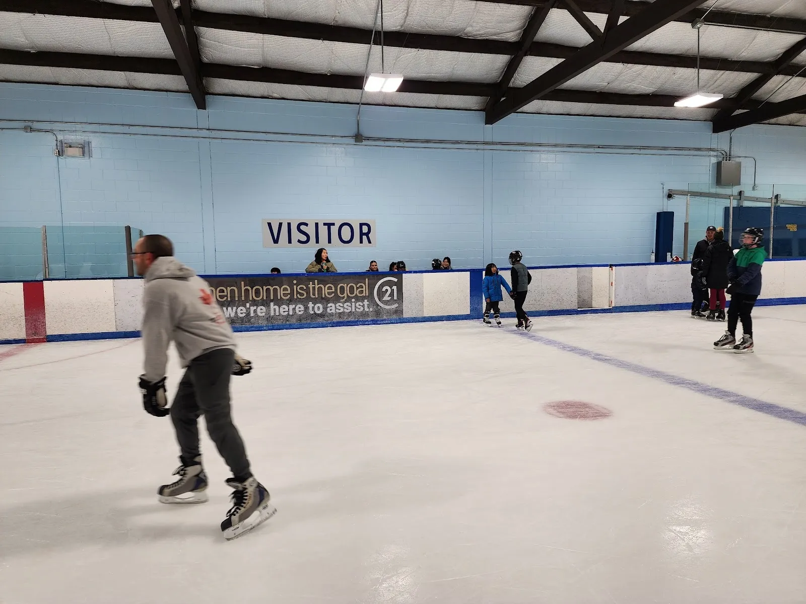 A skater near the boards in a public session on a pale blue-walled sheet, others skating far across the ice beneath the Visitor wall and exposed roof beams. — Jack Staples Arena