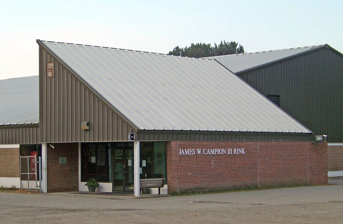 Entrada principal del edificio de la pista con techo metálico gris y una fachada de ladrillo que dice "JAMES W. CAMPION III RINK", vista desde el estacionamiento. — James W. Campion III Rink