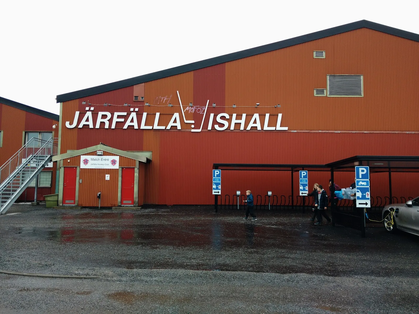 Den röda korrugerade Järfälla Ishall med namnet och logotypen med korslagda klubbor över väggen, sedd från grusparkeringen – Järfälla ishall