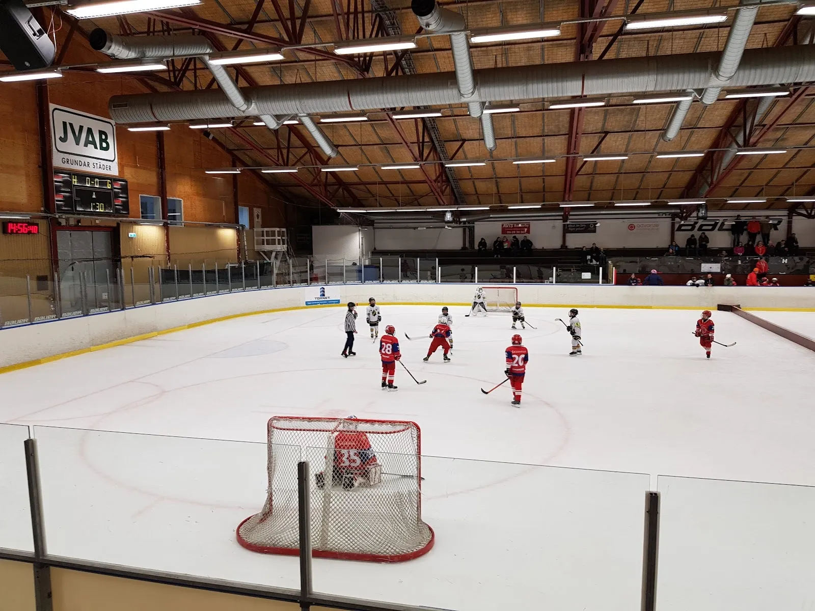 En träning framför ett mål på isen i Järfälla ishall, med trätaket, tegelgaveln och sargreklamen i bakgrunden. – Järfälla ishall