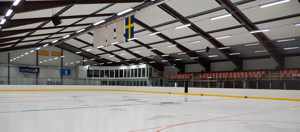 Tom is inne i Järna IP ishall under ett tak med stålfackverk, med Södertälje-skylten på sargen – Järna IP ishall