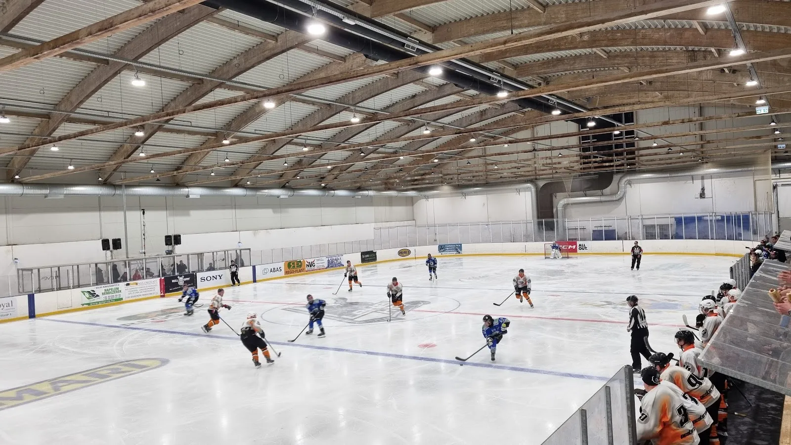 An ice hockey game on the sheet at the Jászberény ice hall, seen from the stand under the arched timber roof — Jászberényi Jégcsarnok