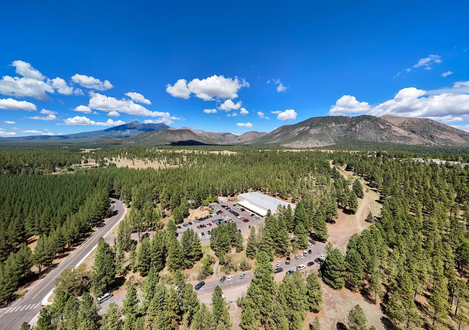 Vista aérea con dron del edificio de hockey del Jay Lively Activity Center, una pista blanca de techo plano rodeada de un bosque de pinos ponderosa con los San Francisco Peaks al fondo y un estacionamiento lleno de autos. — Jay Lively Ice Arena