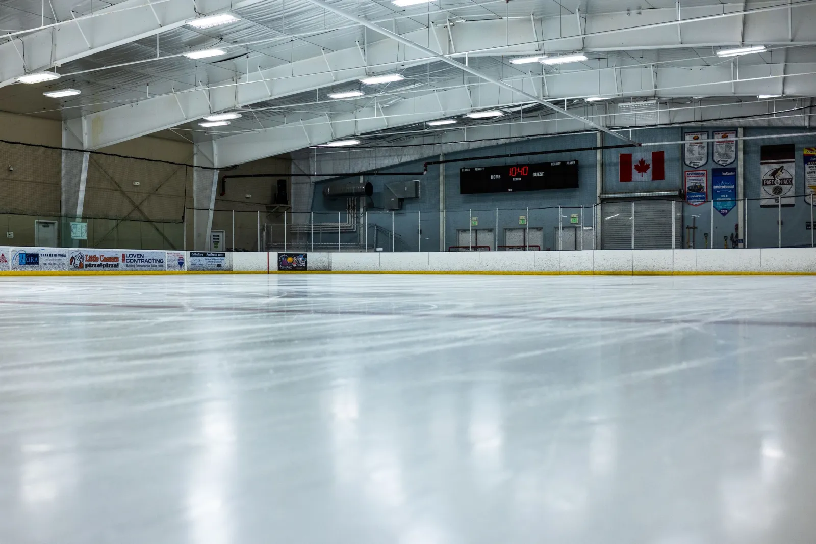 Pista de hielo vacía desde el nivel de la pista hacia las vallas del fondo, con un marcador en '10:40', una bandera de Canadá, mantas de campeonato y una cortina de garaje enrollada en la pared del fondo, y anuncios de negocios locales en las vallas (Little Caesars, OrthoCare, Loven Contracting). — Jay Lively Ice Arena
