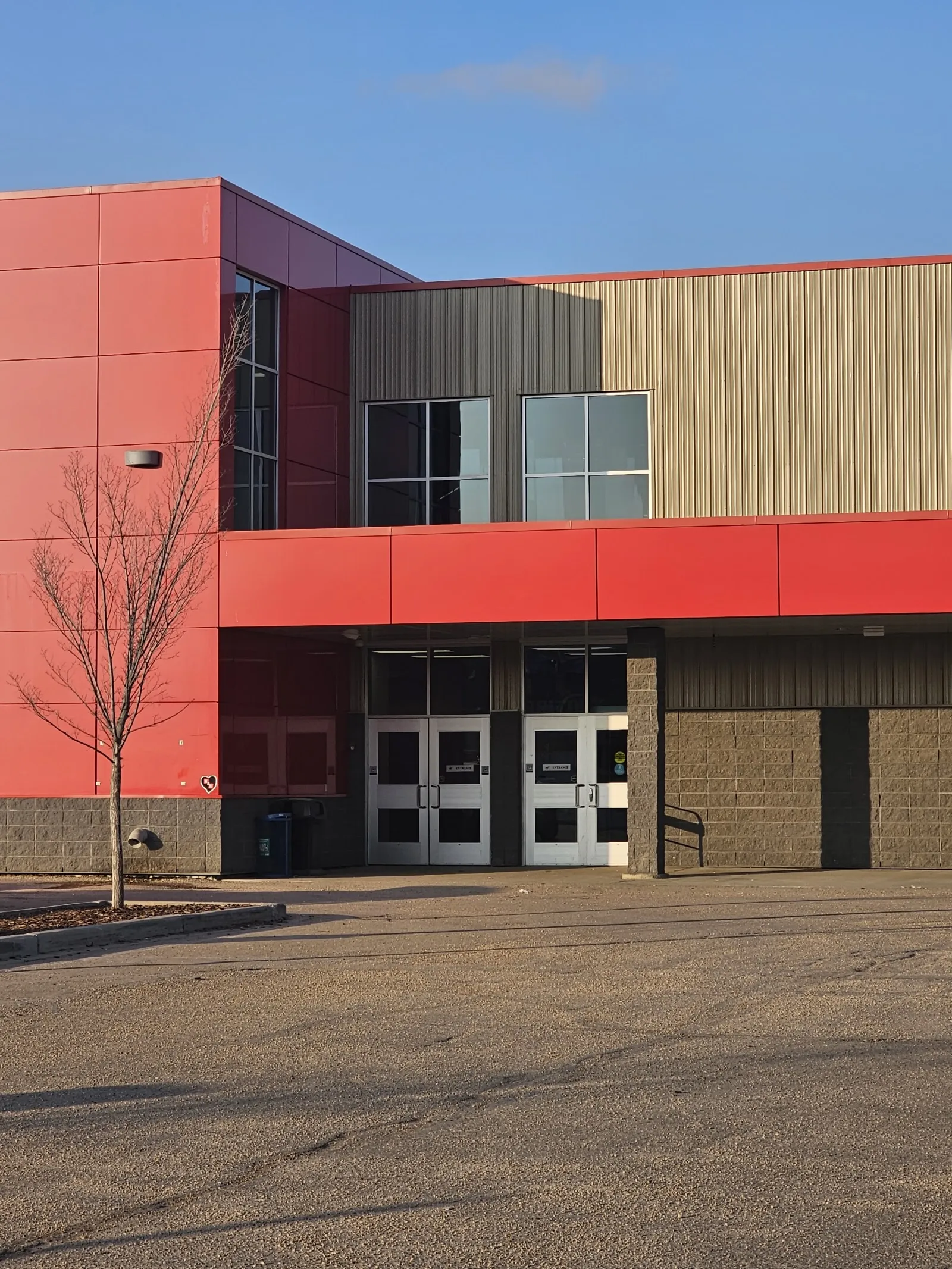 El frente rojo y beige de JDA Place en Whitecourt con sus puertas dobles de entrada, visto al otro lado del estacionamiento — JDA Place
