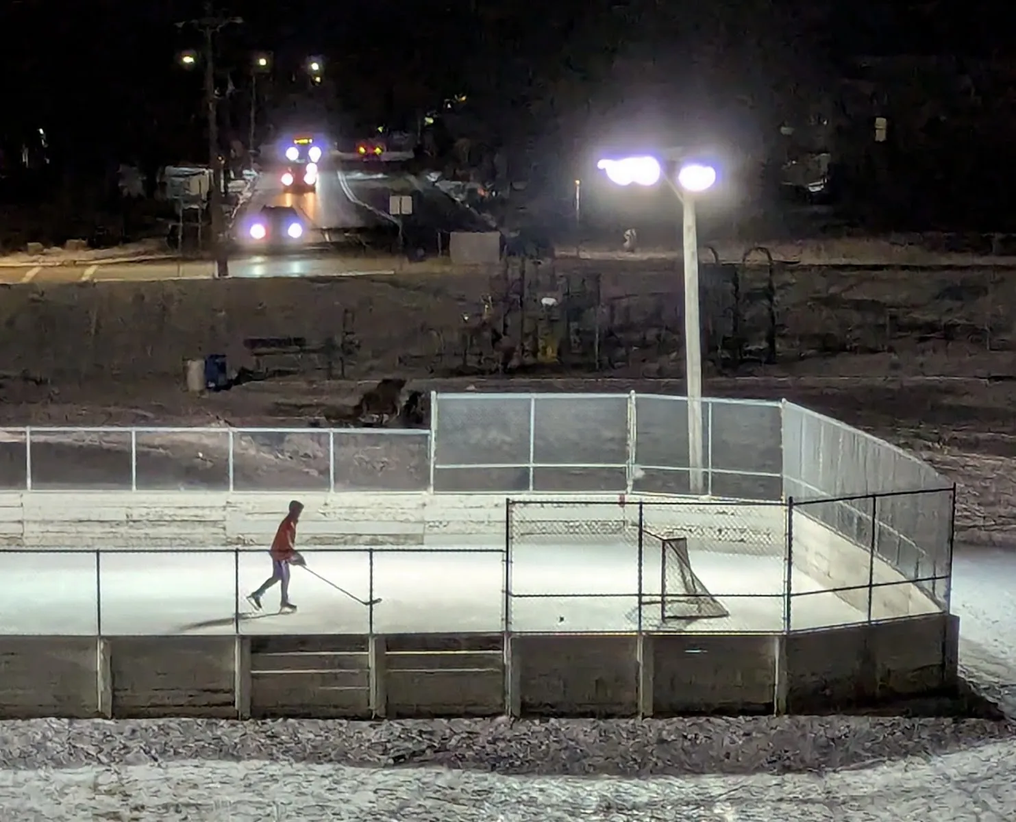 The lit, board-enclosed rink at Jefferson Park at night with one skater and a goal — Jefferson Park Ice Rink