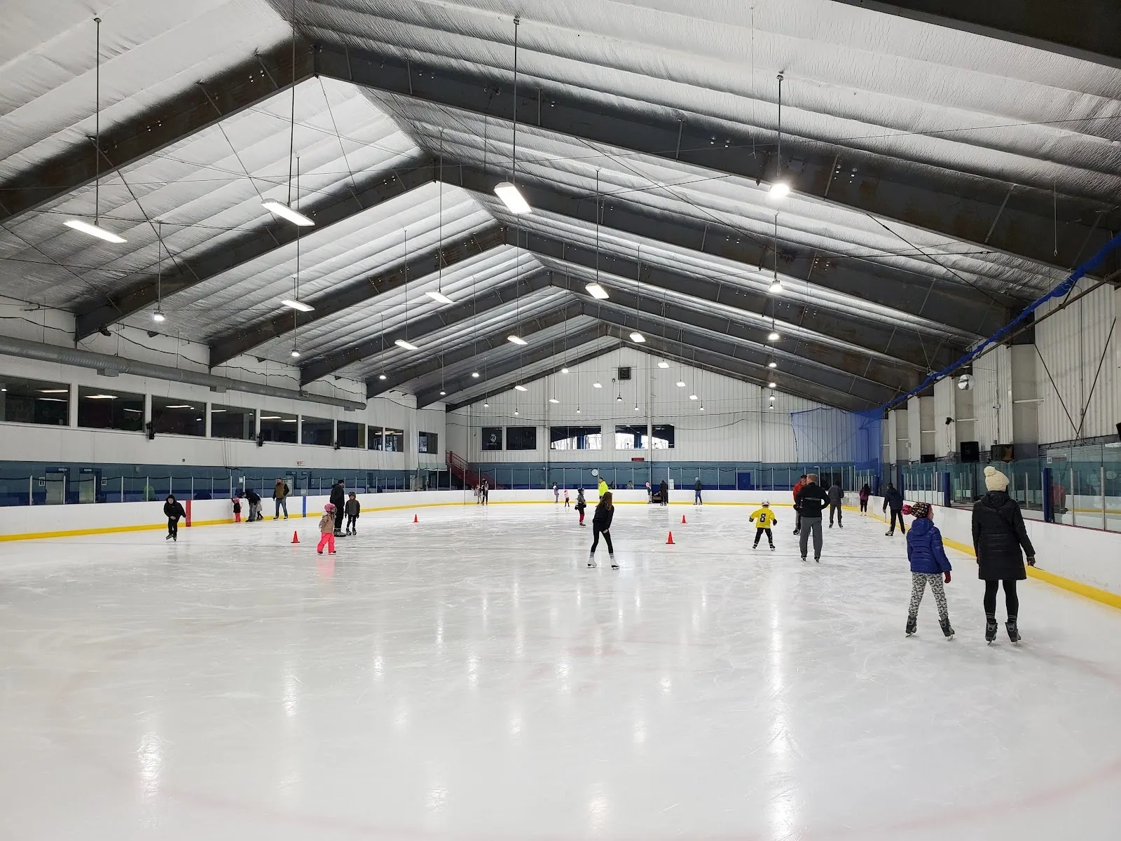 A public session on a wide sheet, skaters spread across white ice under a long gabled roof with steel braces and a blue and red wall behind. — Jersey Shore Arena