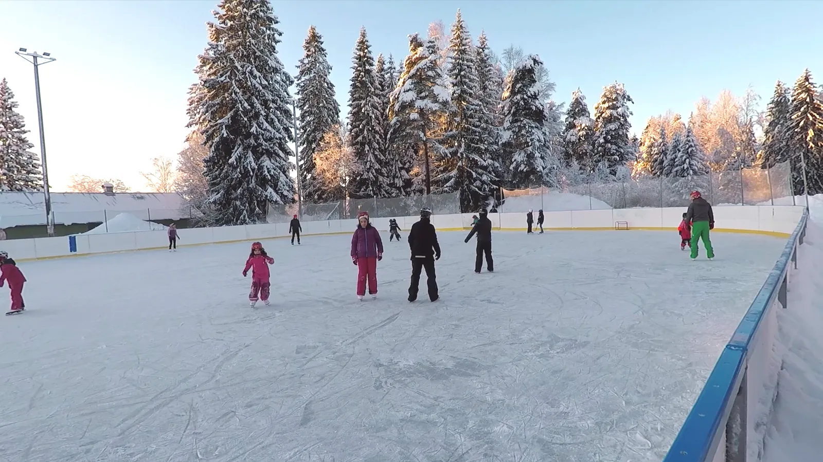 The artificial-ice rink at Jessheim sports park on a bright winter day: children skating in small groups along the boards of the outdoor rink, with snow-laden spruce trees behind. — Jessheim idrettspark kunstisbane