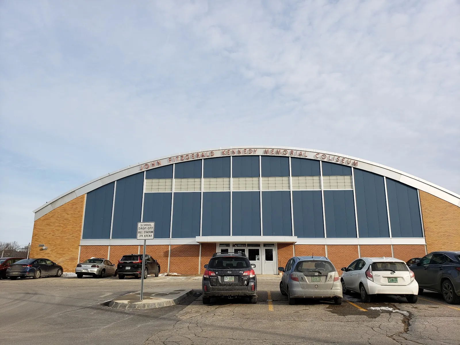 Front of the arched-roof brick building with "JOHN FITZGERALD KENNEDY MEMORIAL COLISEUM" lettering above the entrance and a parking lot in front. — JFK Memorial Coliseum
