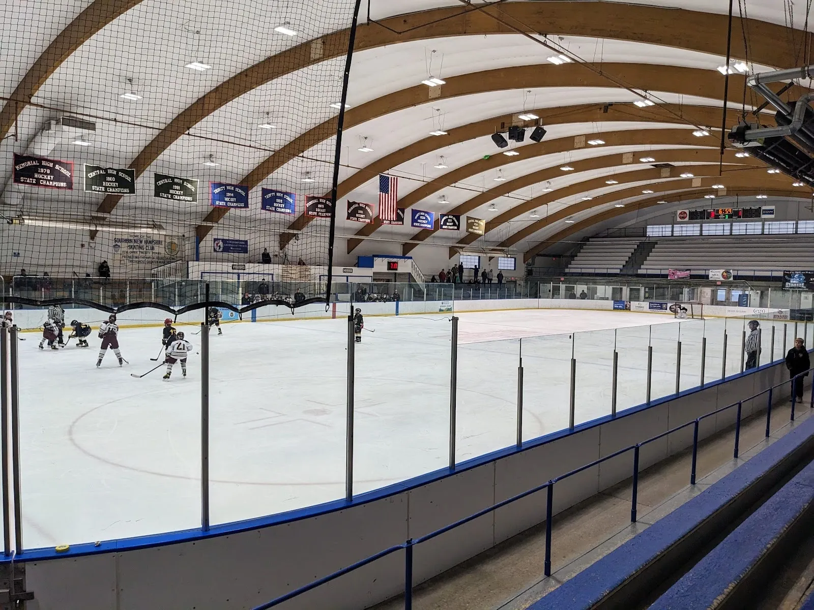 A public session seen over the blue-edged boards, skaters spread across the ice beneath a curved timber roof hung with pennants. — JFK Memorial Coliseum